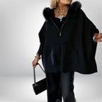 Women’s cape coat in black with fur-trimmed hood, elegant wide silhouette and pockets styled with layered jewelry for chic winter outfits