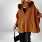 Women’s cape coat in warm brown with fur hood and oversized silhouette, styled with layered jewelry for classic seasonal elegance