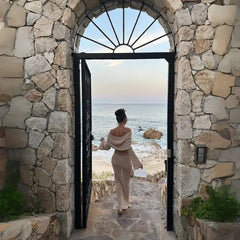 Women's one-shoulder long-sleeve knitted vacation dress in beige walking through stone gate to ocean.