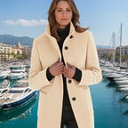 Wool pea coat in cream on a woman posing by a marina with boats and waterfront buildings.