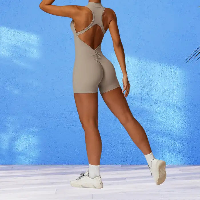 workout romper in neutral beige featuring open back design against vibrant blue wall highlighting its athletic silhouette