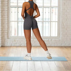 workout romper in charcoal gray with open back design shown in bright studio with white brick walls and blue yoga mat